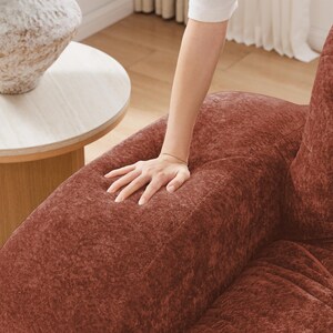 May include: Close-up of a rust-colored, textured sofa. A person's hand is pressing into the plush fabric, highlighting its softness. A small, round side table with a white top and a decorative vase are in the background.