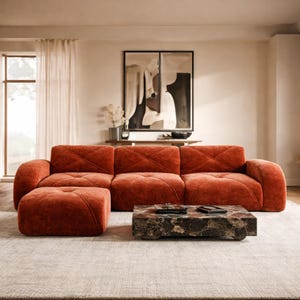 May include: A modern living room setup featuring a rust-colored sectional sofa with a matching ottoman and a square marble coffee table. The sofa has a quilted design. A large abstract artwork hangs on the wall.