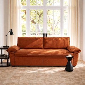 May include: A burnt orange sofa with two large back cushions and two armrests. The sofa is in front of a large window with white trim and sheer curtains. A black side table and lamp are to the left, and a black side table with a candle is to the right.