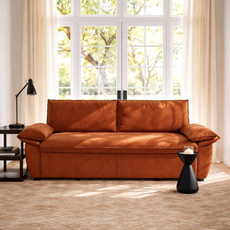 May include: A burnt orange sofa with two large back cushions and two armrests. The sofa is in front of a large window with white trim and sheer curtains. A black side table and lamp are to the left, and a black side table with a candle is to the right.