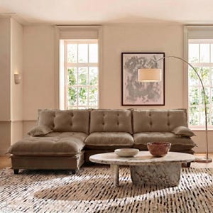 May include: A large, sectional sofa in a neutral beige color with button tufting. A round marble coffee table with a bowl and a woven basket sits on a patterned rug. A floor lamp and artwork complete the living room setting.
