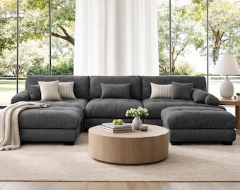 Puede incluir: Un gran sofá seccional gris oscuro con múltiples cojines y una otomana a juego. El salón cuenta con una mesa de centro redonda de madera, una mesa auxiliar con una lámpara y una alfombra de color neutro. Cojines decorativos y una manta añaden un ambiente acogedor.