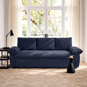 May include: A navy blue sofa with three back cushions and two arm cushions. The sofa is in front of a large window with white curtains. A black side table with a lamp and a small black table with a candle are on either side of the sofa.