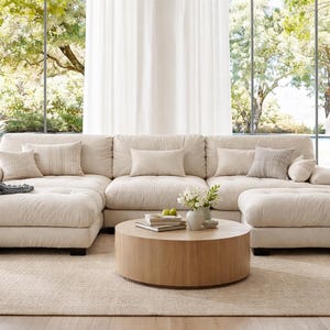 May include: A large, cream-colored sectional sofa with deep cushions and a chaise lounge on each end. The sofa features several matching throw pillows, a textured gray throw blanket draped over one side, and a round, light-wood coffee table.
