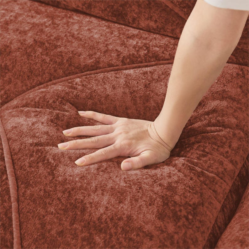 May include: Close-up of a hand pressing into a plush, rust-colored fabric. The fabric has a textured, velvety appearance, with visible stitching along the seams. The image highlights the softness and tactile quality of the material.