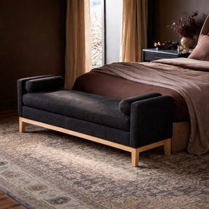 May include: A black upholstered bench with light wood legs and armrests. The bench is placed at the foot of a bed with a brown comforter and a patterned rug. The room has warm lighting and neutral-toned curtains.