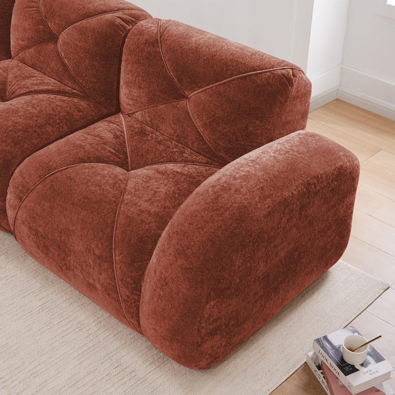 May include: A close-up of a rust-colored, modular sofa with a geometric design. The sofa has a plush, velvety texture and is positioned on a light-colored rug. The armrest is curved and the sofa appears to be in a living room setting.