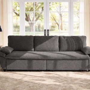 May include: A large, gray sofa with three back cushions and two arm cushions. The sofa is in front of a large window with a view of trees. A black side table and lamp are to the left.