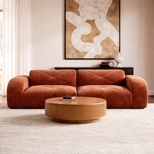 May include: A modern living room setup featuring a rust-colored sofa with a tufted design, a round wooden coffee table, and a large abstract painting. The room is decorated with neutral tones and a patterned rug.