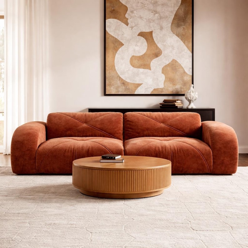 May include: A modern living room setup featuring a rust-colored sofa with a tufted design, a round wooden coffee table, and a large abstract painting. The room is decorated with neutral tones and a patterned rug.