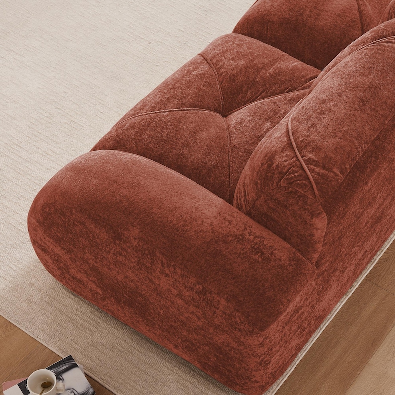May include: Close-up of a rust-colored sofa with a textured, velvety fabric. The sofa features a rounded armrest and tufted back cushions. The cushions have a geometric pattern. The sofa sits on a light-colored rug.
