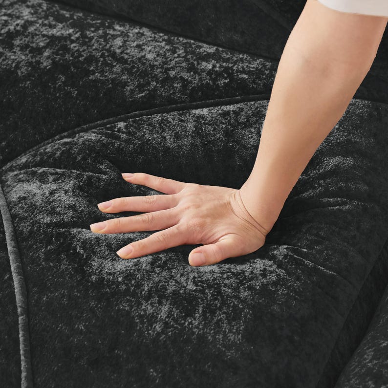 May include: A black velvet sofa cushion with a soft, plush texture. The fabric features a deep, dark color with subtle light-reflecting qualities, showing a tufted or quilted pattern with visible stitching lines across the surface.