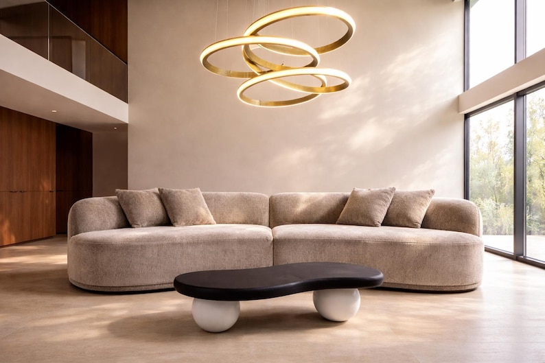 Puede incluir: Un sal&oacute;n moderno con un gran sof&aacute; beige curvo y cojines a juego. Una mesa de centro negra en forma de ri&ntilde;&oacute;n descansa sobre dos bases esf&eacute;ricas blancas. Arriba, una l&aacute;mpara de ara&ntilde;a dorada circular ilumina el espacio.