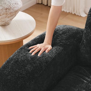 May include: A black armchair featuring a textured, velvet-like fabric upholstery. The chair has a rounded, padded armrest and a deep seat cushion, showcasing a dark, mottled appearance with subtle variations in light and shadow across the surface.