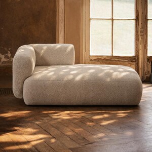 May include: A beige, curved chaise lounge with a rounded armrest. The sofa has a textured, plush fabric. The room has a wooden floor and a window with sunlight streaming through, creating shadows on the furniture.