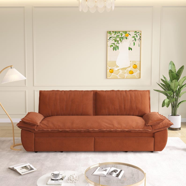 May include: A burnt orange corduroy sofa with two back cushions and two armrest cushions. The sofa is in a living room setting with a painting of a cat and oranges, a floor lamp, and a potted plant.
