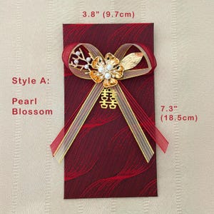 Puede incluir: Un sobre rojo burdeos, de 18,5 cm de largo y 9,7 cm de ancho, adornado con un lazo de cinta dorada y roja. El lazo presenta una flor dorada con perlas y un s&iacute;mbolo de doble felicidad. El texto dice "Style A: Pearl Blossom."