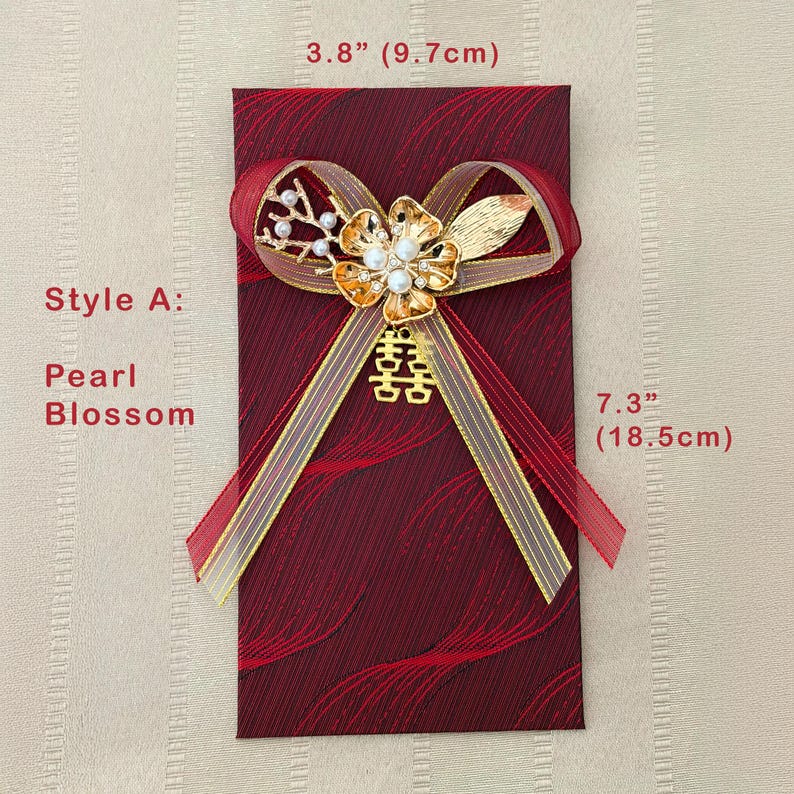 Puede incluir: Un sobre rojo burdeos, de 18,5 cm de largo y 9,7 cm de ancho, adornado con un lazo de cinta dorada y roja. El lazo presenta una flor dorada con perlas y un s&iacute;mbolo de doble felicidad. El texto dice "Style A: Pearl Blossom."