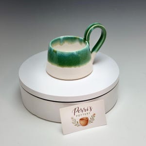 May include: A small, handmade ceramic mug with a white base and a vibrant green upper section. The mug has a curved handle and a glossy finish. A business card with the text "Perri's Pottery" is in the foreground.