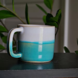 Könnte beinhalten: Eine Keramik-Tasse mit weißem Oberteil und türkisfarbenem Boden, getrennt durch ein dünnes gelbes Band. Die Tasse hat einen gebogenen Henkel und steht auf einer dunklen Oberfläche. Grüne Blätter sind im Hintergrund zu sehen.