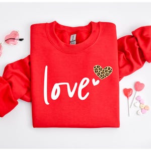 May include: A red sweatshirt with the word "love" in white script. A leopard print heart is on the upper right. Heart-shaped sunglasses and Valentine's Day candy are also in the image.
