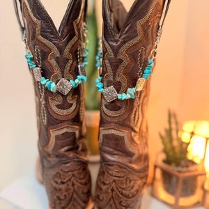 May include: A pair of brown leather cowboy boots with intricate stitching and decorative patterns. Turquoise and silver boot chains add a pop of color and detail. The boots have a square toe and a medium heel.