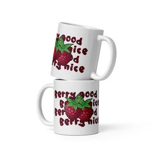 Op de afbeelding: Twee witte keramische mokken op elkaar gestapeld. Elke mok heeft een frambozenillustratie met groene bladeren en de tekst "Berry Good, Nice". De mokken hebben een gebogen handvat.