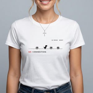Op de afbeelding: Wit T-shirt met een pixelachtige dinosaurus die langs cactussen rent. De tekst "NO CONNECTION" is rood, en "HI 99999 99997" staat erboven. Een zilveren kruis ketting is ook zichtbaar.