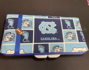Puede incluir: Estuche rectangular de tela con un patrón azul y blanco con la palabra "CAROLINA" y el logotipo de UNC. Se adjuntan una cinta azul y un botón en forma de balón de fútbol. El estuche tiene un cierre blanco.