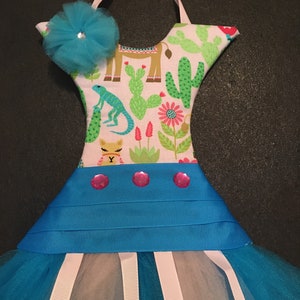 May include: A decorative dress-shaped wall hanging with a turquoise tulle skirt and a bodice featuring a desert-themed print with cacti, llamas, and a lizard. The bodice has a turquoise flower with a rhinestone center. Pink ribbon accents the skirt.