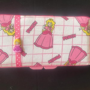May include: A pink and white rectangular case featuring a repeating pattern of Princess Peach from the Super Mario Bros. series. The case has a pink ribbon with a Princess Peach charm and the word "PEACH" printed on the case.