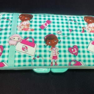 May include: A rectangular, fabric pencil case featuring a green and white checkered pattern. The case is decorated with cartoon images of a doctor, a lamb, hearts, bandages, and stethoscopes. A light green ribbon runs down the center.