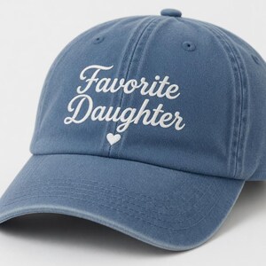 Puede incluir: Gorra de b&eacute;isbol azul con las palabras "Favorite Daughter" en escritura blanca y un peque&ntilde;o coraz&oacute;n blanco. La gorra tiene una visera curva y un bot&oacute;n en la parte superior.