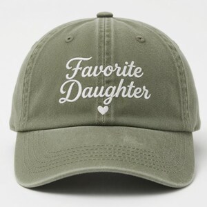 Puede incluir: Gorra de b&eacute;isbol verde oliva con la frase "Favorite Daughter" en escritura blanca, con un peque&ntilde;o coraz&oacute;n blanco. La gorra tiene una visera curva y un cierre de tela.