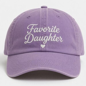 Puede incluir: Gorra de b&eacute;isbol lavanda con la inscripci&oacute;n bordada "Favorite Daughter" en blanco y un peque&ntilde;o coraz&oacute;n blanco. La gorra tiene una visera curva y un cierre de correa de tela.