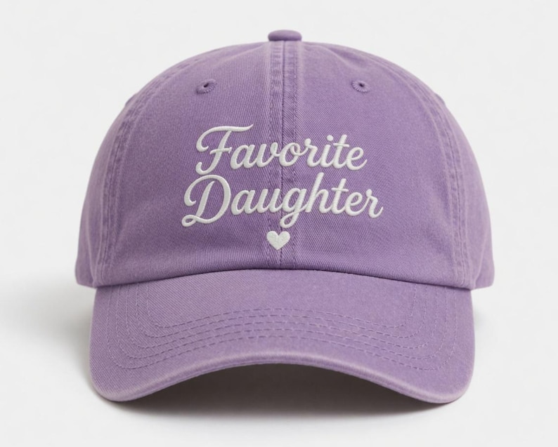 Puede incluir: Gorra de b&eacute;isbol lavanda con la inscripci&oacute;n bordada "Favorite Daughter" en blanco y un peque&ntilde;o coraz&oacute;n blanco. La gorra tiene una visera curva y un cierre de correa de tela.