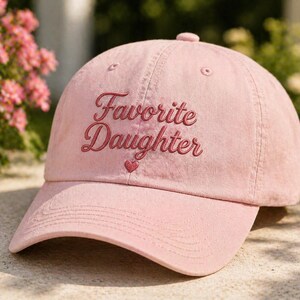 Puede incluir: Gorra de b&eacute;isbol rosa claro con la frase bordada "Favorite Daughter" en cursiva. Un peque&ntilde;o coraz&oacute;n est&aacute; bordado debajo del texto. La gorra tiene una visera curva y un bot&oacute;n en la parte superior.