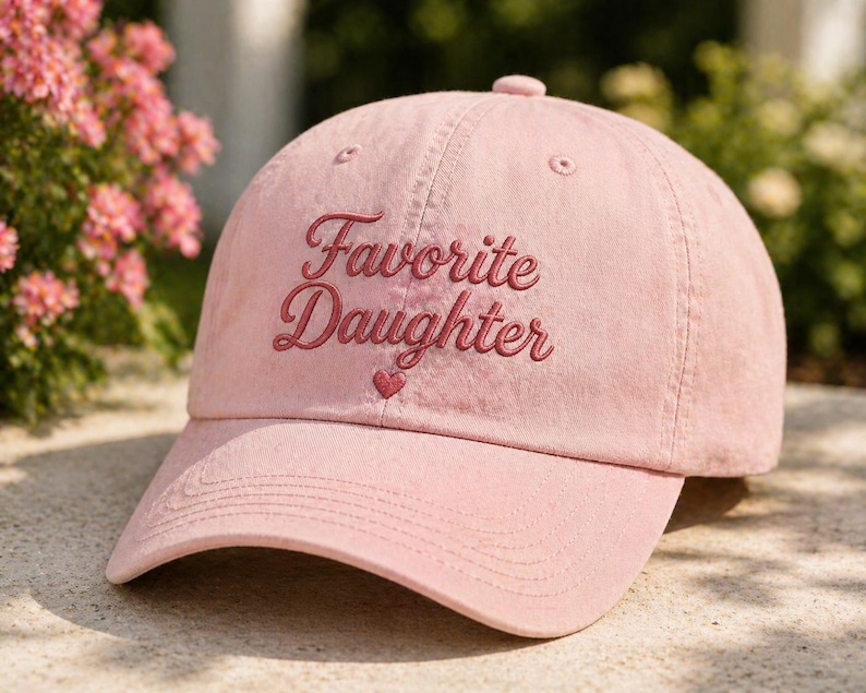 Puede incluir: Gorra de b&eacute;isbol rosa claro con la frase bordada "Favorite Daughter" en cursiva. Un peque&ntilde;o coraz&oacute;n est&aacute; bordado debajo del texto. La gorra tiene una visera curva y un bot&oacute;n en la parte superior.
