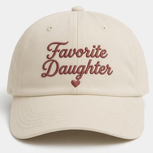 Puede incluir: Gorra de b&eacute;isbol color crema con el texto bordado "Favorite Daughter" en fuente burdeos. Un peque&ntilde;o coraz&oacute;n burdeos est&aacute; bordado debajo del texto. La gorra tiene una visera curva y una correa ajustable.