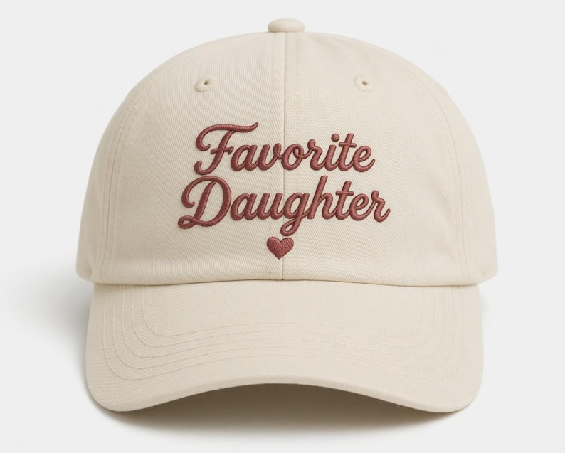 Puede incluir: Gorra de b&eacute;isbol color crema con el texto bordado "Favorite Daughter" en fuente burdeos. Un peque&ntilde;o coraz&oacute;n burdeos est&aacute; bordado debajo del texto. La gorra tiene una visera curva y una correa ajustable.