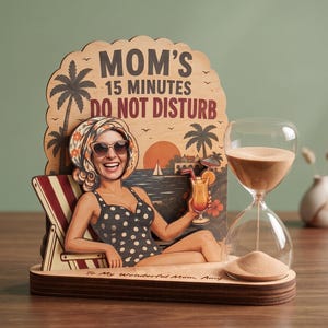 Puede incluir: Un letrero de escritorio de madera con la ilustración de una persona en traje de baño de lunares y pañuelo, descansando en una silla con un cóctel. El texto dice MOM'S 15 MINUTES DO NOT DISTURB y To My Wonderful Mom, Amy. Un reloj de arena de cristal está al lado.