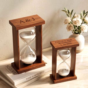 May include: Two rectangular wooden hourglasses with white sand. The larger hourglass features the engraved text A & J on the top frame. The smaller hourglass features the engraved text Forever is made of moments. 1. 24.2026 on the top frame.