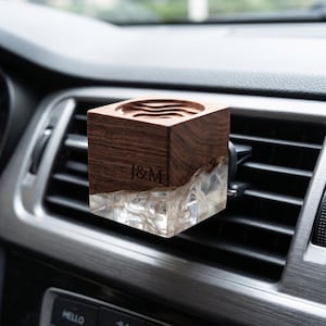 Puede incluir: Un clip ambientador de coche cuadrado con una parte superior de madera marrón oscura y una base de resina transparente con patrones blancos en espiral. La sección de madera está grabada con las letras J&M y la superficie superior tiene un corte circular.