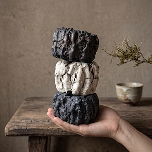 May include: A stack of three small, cylindrical ceramic vessels with highly textured, rugged surfaces. The top and bottom vessels are dark charcoal black, while the middle vessel is a contrasting off-white with deep, irregular vertical ridges.