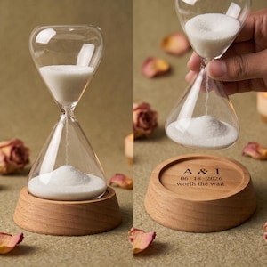 May include: A clear glass hourglass filled with white sand, resting on a round wooden base. The base is engraved with the text A & J, 06 18 2026, and worth the wait. The hourglass is shown both standing on the base and being held above it.