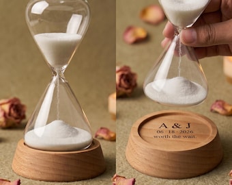 Puede incluir: Un reloj de arena de cristal transparente lleno de arena blanca, sobre una base redonda de madera. La base tiene grabado el texto A & J, 06 18 2026, y worth the wait. El reloj de arena se muestra tanto sobre la base como suspendido.