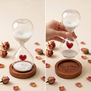 May include: A glass hourglass containing white sand and a small red heart-shaped glass element. The hourglass rests on a circular wooden base engraved with the text Liam & Olivia, Forever Yours, 2024.10.12. Dried rose petals are scattered around.