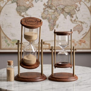 May include: Two rotating hourglasses with dark wood frames and brass-toned metal supports. The larger hourglass features the engraved text M&J on the top wooden cap and contains tan sand. A small glass vial with a cork stopper filled with sand sits nearby.