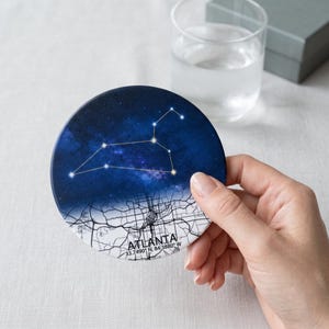 May include: A circular coaster featuring a deep blue night sky with a white constellation pattern on the top half and a black and white street map of Atlanta, Georgia, on the bottom half, including the text ATLANTA 33.7490 N, 84.3880 W.
