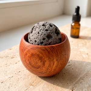May include: A spherical, porous gray lava stone sits inside a small, smooth, reddish-brown wooden bowl. Beside the bowl stands a small amber glass dropper bottle with a black cap, containing a liquid, set against a bright, neutral background.